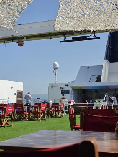 The image depicts an outdoor deck area of a ferry or cruise ship, under a camouflage netting canopy, on a bright, sunny day. The deck is covered with artificial turf and furnished with numerous wooden tables and red chairs, many of which bear the "ESTRELLA DAMM" logo. In the background, a white ferry structure rises, with a satellite dish visible atop it. A man in a floral shirt and hat stands on the deck, facing away from the camera, appearing to interact with an object on a table. Further in the background, other passengers are seated at tables in a shaded area. The distant horizon shows a hazy outline of hills or mountains. The location is implied to be the city of Magaluf, Spain, suggested by the presence of "ESTRELLA DAMM," a Spanish beer brand popular in tourist areas.