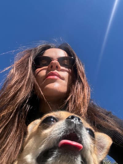 The image presents a low-angle, upward-facing outdoor portrait, taken in Alcoi / Alcoy, Spain. The upper half of an adult female's face and her upper torso occupy the upper portion of the frame. She has long, dark brown hair, and is wearing dark sunglasses with thin frames. Her lips are a reddish-pink color. Strong direct sunlight casts shadows under her nose and chin. She appears to be wearing a dark, possibly black, ribbed garment. In the foreground, positioned below the female and closer to the camera, is the head and upper chest of a small dog, consistent with a Chihuahua or similar breed, with light brown (tan/fawn) fur. The dog's mouth is slightly open, revealing a pink tongue partially extended, and its wide eyes are looking upwards. Both subjects are intensely illuminated by direct sunlight. The background is a clear, bright blue sky, featuring a distinct sun flare in the upper right quadrant, indicating a sunny day. Minimal out-of-focus green foliage is faintly visible at the very bottom right edge of the frame, behind the dog.