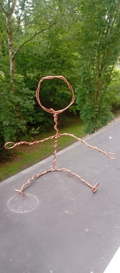 An abstract wire sculpture resembling a human figure is positioned on a dark, textured surface, possibly a railing or ledge, with a lush green, leafy background suggesting an outdoor, possibly urban, setting. The sculpture, made of twisted copper-colored wire, is abstractly formed with a circular head and a body, limbs, and feet fashioned from bent and coiled wire. The surface it rests on has a circular white mark and some scattered lighter speckles. Behind the sculpture, the foliage is dense and vibrant green, with hints of tree trunks visible. The overall impression is one of a simple, artistic creation placed outdoors. The image does not contain specific landmarks to place it in Karviná, Czechia.