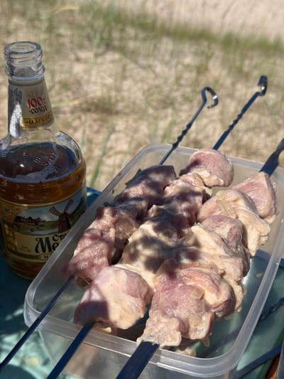 This media file shows skewers of raw meat in a clear plastic container, placed on a greenish surface next to a bottle of alcohol. Two metal skewers are visible, one with meat threaded onto it, the other empty and lying on the sand. The meat appears to be marinated, with a creamy sauce coating the pieces. The bottle has a label with text and an illustration of a windmill. Part of the label reads "100% СОЛОД" (100% Malt) and "Мягкое" (Soft). Other visible text on the label includes what appears to be Latvian. The background consists of sand and blurred green grass, suggesting an outdoor setting, likely a beach, given the location context of Plieņciems, Latvia. The lighting indicates it is daytime, with sunlight casting shadows. The overall scene evokes a sense of preparation for a barbecue or outdoor meal. There are no people visible in the image.