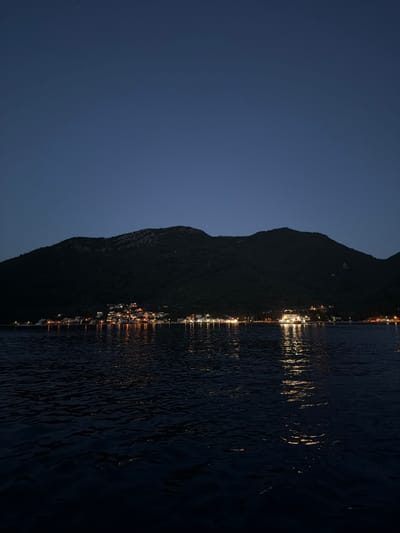 The image shows a nighttime view of a coastal town nestled at the base of a dark, tree-covered mountain. The town, likely located in or near Đurići, Montenegro, is illuminated by numerous lights, suggesting a settlement along the shore. The lights reflect on the dark, rippling surface of the water in the foreground, creating streaks of golden illumination. The sky above is a deep, dark blue, with a faint star visible in the upper left quadrant. The overall scene depicts a tranquil, dimly lit landscape with human habitation against a natural backdrop.