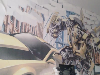 The image depicts a mural or wallpaper featuring a stylized scene with a yellow car in the foreground and a robotic figure in the background, possibly a transformer character. The car's front is visible, with a dark windshield and hood. Behind the car, there is a large, intricately detailed robot with yellow and gray metallic components. The background includes elements that resemble buildings, with brick patterns and architectural details, along with industrial-looking structures and scaffolding. A small, white robot with a black visor is positioned near the bottom right of the yellow car. The overall scene is dynamic and likely intended as a decorative element. The prompt's mention of "Мамлют қаласының әкімшілігі, Kazakhstan" is not visually represented in the image and appears to be an irrelevant contextual addition.