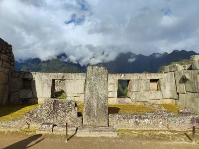 Ancient stone ruins sit nestled against a backdrop of lush green mountains partially obscured by swirling clouds. The stonework is gray and weathered, marked by age and moss. There are several windows in the ruins, offering glimpses of the landscape beyond. A monolithic stone pillar stands prominently in the center. The ground in front of the structure is a mixture of grass and dirt.