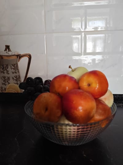 The image depicts a still life composition featuring a bowl of fruit and a decorative pitcher on a dark countertop. In the foreground, a clear glass bowl contains a variety of fruits, including several nectarines or peaches with red and orange hues, and a pale green apple. Behind the bowl, a bunch of dark grapes is visible. To the left, a decorative pitcher with a white base adorned with a gold pattern and a gold handle stands near a slice of lemon. The background is a white tiled wall, with a slight reflection of light visible on the tiles. The scene is set in what appears to be a kitchen environment. The image does not provide any information about the city of Kogon City, Uzbekistan.