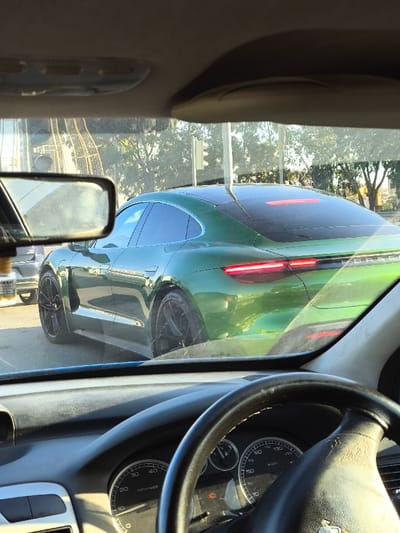 The image depicts an urban traffic scene in Gonyeli, Northern Cyprus, observed from the interior of a stationary vehicle during daylight hours.

The primary subject is a dark green Porsche Taycan electric sedan, viewed from its rear-right quarter, positioned in the lane ahead of the observer. The Taycan features black multi-spoke wheels and illuminated red LED taillights, including a full-width light bar and main light elements. The "PORSCHE" lettering is visible across the rear fascia. Directly behind the Taycan, a portion of another dark-colored vehicle is visible.

In the foreground, the observer's vehicle interior is visible, including the top of a dark dashboard, a black, worn steering wheel, and an instrument cluster. The speedometer reads 0 km/h and the tachometer indicates 0 rpm, with an amber warning light illuminated. A rear-view mirror on the left side of the frame reflects ambiguous interior elements. A small, clear bottle with a white label bearing the Turkish word "DIKKAT" (CAUTION) hangs from the rear-view mirror.

The background shows green trees, a traffic light pole with a white signal box, and a prominent, large decorative metal structure characterized by a curved, lattice-like design, situated to the far left. The overall environment suggests an urban roadway or intersection.