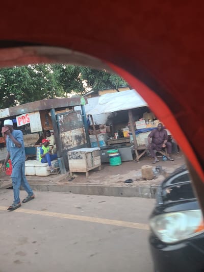 This image depicts a street scene in Karu, Nigeria. The foreground is dominated by the interior of a vehicle, likely a tricycle or motorcycle taxi, with a red canopy partially obscuring the view. Beyond this, a market stall is visible.
Several people are present. In the middle ground, a man in a dark purple traditional outfit is seated on a wooden bench outside a stall. To his right, a large green barrel is situated on the ground. Further down the stall, two individuals are seated, one wearing a blue shirt and yellow vest, and another in a red cap, both appearing to be interacting. In the foreground, a man dressed in a light blue traditional outfit and a white cap is standing and appears to be looking down or to the side, with his hand near his face. He is holding a red and green object, possibly a box or package, in his other hand. A sign with the text "POS" is visible on the stall.
The setting appears to be a bustling outdoor market or street-side business area. The ground is dirt or unpaved, with some debris scattered around. The stalls are made of wood and corrugated metal, suggesting a somewhat informal setup. The lighting suggests it is daytime, likely late morning or afternoon, with the sun casting shadows. The overall atmosphere seems ordinary, with people engaged in their daily activities. The image is taken from a mobile device, with a slight blur, possibly due to movement.