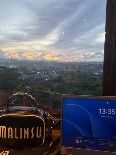 The image presents an elevated view of a cityscape at what appears to be sunset or dusk, originating from an indoor vantage point through a window in Benoa, Indonesia. In the foreground, positioned on the left, is a black handbag with "MALINSU" printed in white capital letters on its front, featuring a studded top handle and a silver chain strap. To the right of the handbag, the visible portion of a laptop screen displays a blue graphical interface. The screen shows a digital clock reading "13:35," the date "пятница, 20 марта" (Friday, March 20), and a weather forecast for "Санкт-Петербург" (Saint Petersburg) indicating 5°C. A "1080" resolution sticker is affixed to the bottom left of the laptop's screen bezel.

The midground reveals an expansive urban landscape characterized by a dense arrangement of residential buildings, most exhibiting tiled roofs, interspersed with abundant green foliage and trees. Various artificial lights are visible across the cityscape, suggesting evening conditions. The background features a cloudy sky exhibiting sunset hues, transitioning from vibrant orange and yellow near the horizon to shades of grey and purple in the upper portions. The visible sunset or dusk lighting is inconsistent with the 13:35 time displayed on the laptop screen.