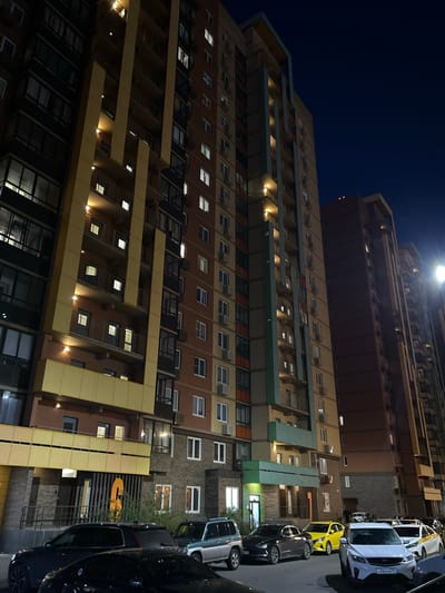A low-angle, nighttime photograph captures a modern, multi-story residential building in the city of Mirny, Russia. The building's facade is a mix of red brick and lighter colored panels, with numerous balconies and windows. Many of the windows are illuminated, suggesting occupancy. A bright yellow car is parked in front of the building, alongside other vehicles including a dark SUV and a white sedan. The sky is dark blue, indicating it is nighttime, and a street light is visible on the right side of the frame, casting a bright light onto the scene. The overall impression is one of urban nighttime ambiance.