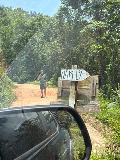 The image, taken from inside a vehicle, shows a dirt road in Hoàng Su Phì Commune, Vietnam. A weathered wooden signpost, marked "NAM LY," stands to the right of the road. A man in a green shirt and shorts is standing on the road near the signpost. The environment is lush with green vegetation, indicating a rural or natural setting. The vehicle's side mirror is visible in the foreground.