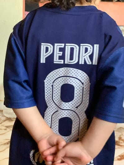 A child, viewed from the back in Sidi Slimane Moul Al Kifane, Morocco, wears a dark blue (navy) sports jersey. The name "PEDRI" is printed in large, white, double-lined block letters across the upper back. Below the name, the number "8" is prominently displayed in a matching white, double-lined style, filled with a pattern of small dark dots. The child's hands are clasped behind their back, fingers interlocked, at the lower center of the frame. A small white emblem, possibly a crest or laurel wreath, is visible on the lower left side of the jersey. The background features a pale yellow wall to the viewer's left and a blurred reddish-orange surface to the right. The lower portion of the image reveals a light-colored, patterned floor, likely tiled.
