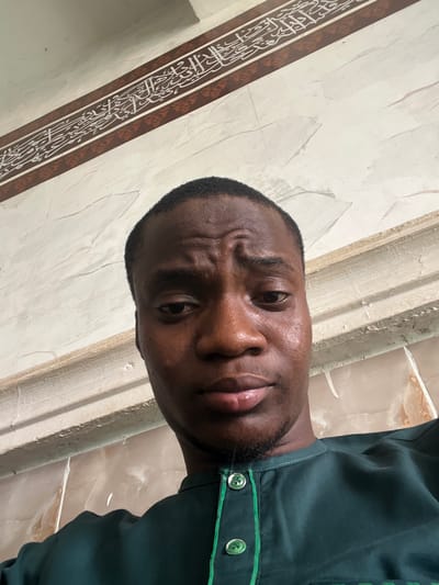 A selfie-style image shows a dark-skinned man with a close-cropped haircut. He is looking directly into the camera with a slight frown. He wears a dark green shirt with a light green trim and two visible silver buttons. The background is a wall with a light beige and white marble-like pattern. A decorative band with brown borders and Arabic calligraphy is visible on the left side of the image. The location is in Katcha, Nigeria.