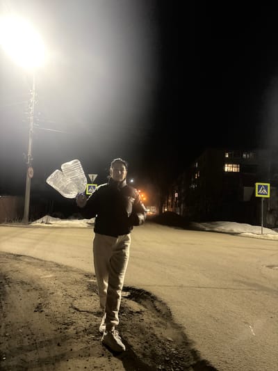 The image depicts an outdoor urban scene at night in Kurbaki, Russia, under artificial illumination. An adult female is positioned in the mid-ground, slightly left of center, facing forward with a direct gaze. She wears a dark, possibly black, puffer jacket, light-colored trousers, and white athletic shoes. In her left hand, she holds a large, clear, visibly crumpled or distorted plastic bottle.
To the immediate left of the individual, a bright street light mounted on a utility pole illuminates the foreground and a portion of the road. A circular speed limit sign displaying '40' is affixed to this pole. The ground comprises a paved road surface, appearing somewhat dirty or gravelly, with a rough dirt and gravel shoulder along its edge. Patches of snow are visible on the shoulders, particularly along the right side of the road.
In the background to the right, a multi-story building, likely an apartment complex, stands with several windows illuminated from within. Further down the road on the right, a yellow square sign with a blue border and a white pedestrian symbol (pedestrian crossing sign) is erected on a pole. The sky above is dark, indicating nighttime conditions. No other individuals or significant actions are depicted.