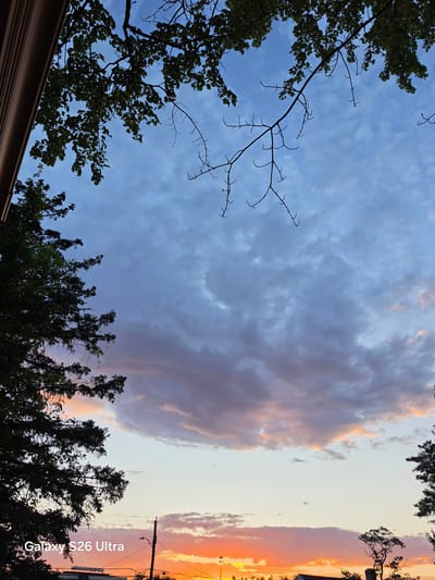 The image depicts a sky with dramatic clouds at dusk, likely taken from West Hartford, United States of America. The sky is filled with various shades of blue, purple, and orange, suggesting the sun is setting or has just set. Silhouettes of trees and branches frame the upper and left sides of the image. In the lower part of the image, telephone poles and wires are visible against the colorful horizon, along with the dark outlines of buildings and more trees. The text "Galaxy S26 Ultra" is superimposed in the lower-left corner of the image.