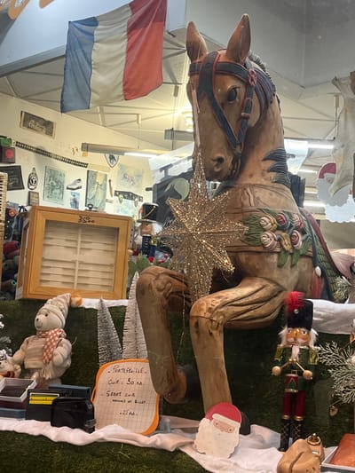 A tilted view captures a carousel horse with festive decorations. A sparkling star adorns its side, accompanied by a nutcracker and a Santa face. A French flag hangs nearby. Beneath the horse, a display includes a glass cabinet, a stuffed bear, miniature Christmas trees, and a price list. Various other items are also visible.