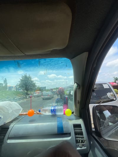 The image is taken from inside a vehicle, looking out through the windshield. The sky is blue with scattered white clouds. Outside, there is moderate traffic on a paved road. Several vehicles, including cars and vans, are visible, as well as motorcycles and pedestrians. On the side of the road, there are buildings and trees. The vehicle's dashboard is in the foreground, with a plastic bag, a bottle of water, and two decorative balls hanging from the rearview mirror. The side mirror of the vehicle is also visible, reflecting a person wearing a white shirt. The location is identified as Gbongan, Nigeria.
