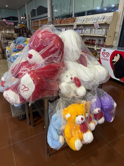 The image depicts a retail display of stuffed teddy bears inside a store. Multiple teddy bears of various sizes and colors, including red, white, blue, yellow, and purple, are prominently displayed, most of them individually packaged in clear plastic. Some of the larger red bears appear to have text and heart designs on them. A sign for "ALGIDA" ice cream is visible on the right side of the frame. The setting appears to be a shop with shelves stocked with various goods in the background, suggesting a general store or a market. The floor is tiled in a reddish-brown color. The scene is captured from a slightly elevated angle, looking down upon the stuffed toys.