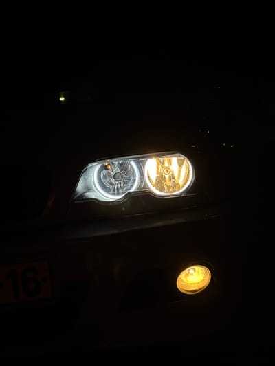 The image shows a vehicle's front end at night. Two illuminated headlights are visible, one with a dual-bulb design where the upper bulb emits a yellow light and the lower one emits a white light. To the right of the dual headlight, another single yellow light is visible. The vehicle itself is black, with a partially visible license plate in the lower right corner with the number 16.