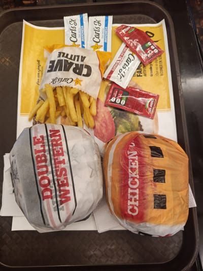 A fast-food meal from Carl's Jr. is displayed on a dark, textured tray. The meal includes two wrapped burgers, a bag of french fries, and several condiment packets.

One burger, wrapped in white paper with red and black stripes, has the text "DOUBLE WESTERN" prominently visible. The second burger, wrapped in orange and clear plastic, shows the word "CHICKEN" in red text. A white paper bag, branded "Carl's Jr." and bearing the phrase "CRAVE A LITTLE," holds a generous serving of golden french fries.

Accompanying the meal are various condiment packets. Two white and blue packets, labeled "Carl's Jr." and "MAYONESA" (mayonnaise), each indicate a net content of 8g. Two red and white packets are visible, one clearly showing "Carl's Jr." and "SALSA DE TOMATE" (tomato sauce/ketchup), also with an 8g net content. Additionally, two smaller red packets, branded "LOS ANDES," feature nutritional warnings such as "ALTO AZÚCAR" (high sugar), "ALTO," and "BAJO."

The tray is lined with a yellow paper featuring Carl's Jr. branding and Spanish text. Partially visible phrases include "Pan com a ajonjoli" (bread with sesame), "la perfección" (the perfection), "FAM..." (likely part of "FAMILY"), "WITH CH," "Grande y," and "¡Que no se diga A COMER EN SER" (Let it not be said... to eat and be served). The Spanish text on the packaging and condiments, combined with the provided context, suggests the location is Samborondón, Ecuador. The image captures a ready-to-eat fast-food order.