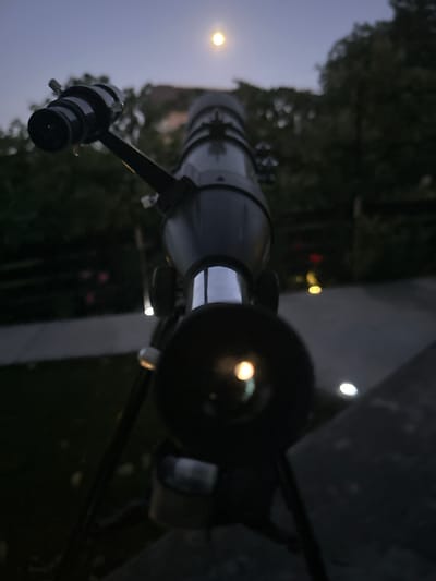 The image shows a telescope aimed at the moon in the twilight sky. The telescope is black and mounted on a tripod. In the background, there are trees and some lights. The scene appears to be in a residential area, possibly a backyard. There are no people visible in the image. The image is a close-up shot of the telescope, with the moon visible in the upper part of the frame. The lower part of the image is blurred, showing the details of the telescope's eyepiece and the surrounding environment. The lighting suggests that it is evening or early morning. The location in the city of Târgu Neamț, Romania, is not discernible from the image itself.