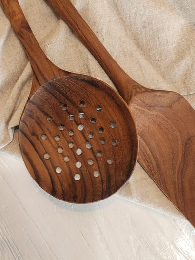 Two wooden cooking utensils, a slotted spoon and a spatula, lie on a piece of light-colored, woven fabric. The slotted spoon, made of dark wood with visible grain, is positioned in the foreground and angled to the left. It features numerous circular perforations across its surface. The spatula, also made of wood with a similar grain pattern, is placed behind and to the right of the spoon, its flat, angled head partially visible. The fabric is laid out on a white, horizontally-lined surface, possibly a table or countertop. The image is a close-up shot, with the lighting casting soft shadows and highlighting the textures of the wood and fabric. The scene appears to be indoors, with no discernible city or geographical context from Maitland, United States of America.