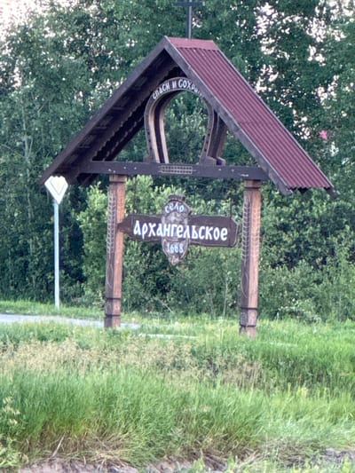 The image shows a wooden signpost in the village of Arkhangelskoye, Russia. The signpost is located in a grassy area with trees in the background. The sign itself is brown with white lettering and depicts the name of the village, "Архангельское," and the year "1668." Above the village name, there is text that reads "СПАСИ И СОХРАНИ," which translates to "Save and Protect." The signpost is supported by two wooden posts carved with decorative patterns. A white diamond-shaped sign is visible on a pole to the left of the main signpost. The overall scene suggests a rural or historical setting.