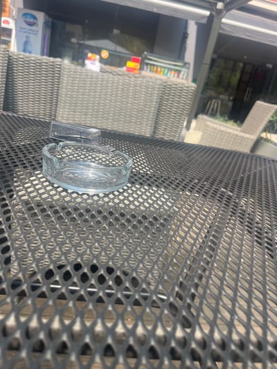 The image shows an outdoor seating area of a cafe or restaurant in Byala Slatina, Bulgaria. In the foreground, on a black metal mesh table, is a clear glass ashtray and a clear plastic lighter. In the background, there are gray wicker chairs and tables, and a building with awnings and shelves displaying bottles and boxes. Sunlight is casting shadows on the table, suggesting it is daytime. The overall scene appears to be a typical urban outdoor dining setting.