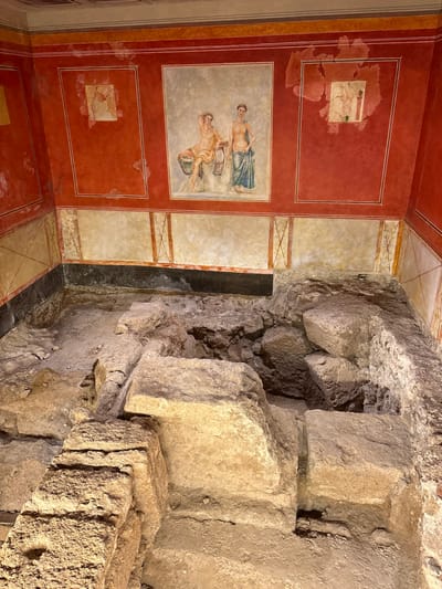 This image captures an archaeological excavation site within a room in Cádiz, Spain. The upper section of the room features walls adorned with preserved frescoes. The dominant upper wall color is a deep reddish-orange, beneath a light-colored ceiling. A large central fresco on the back wall depicts two female figures: one seated or reclining, partially draped, and holding an object; the other standing, partially draped in blue fabric. Two smaller, square-framed frescoes, partially damaged, are visible on the upper left and right walls, each depicting a single figure in motion. A horizontal black band separates the reddish-orange upper wall from a lower, cream-colored section decorated with painted architectural motifs and linear patterns. Visible deterioration, including paint loss and flaking, is present on the frescoes and surrounding wall surfaces.

The lower half of the image showcases excavated ancient foundations composed of irregular, rough-hewn stone blocks and earthen fill. These light-colored stones, varying in size and shape, are stacked to form a multi-level structural base. The excavation extends downwards, revealing layers of historical construction. The scene is illuminated, creating shadows that emphasize the texture and depth of the stone structures. No individuals or modern objects are present.