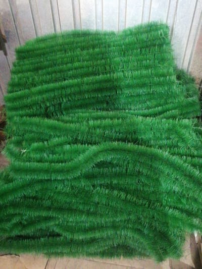 The image depicts a large quantity of green artificial grass, likely a roll of turf, laid out in multiple layers. The material is tightly packed and appears to be synthetic, with individual strands of green fibers visible. The background consists of a ribbed, metallic surface, possibly corrugated metal sheeting. The lighting suggests an indoor environment. There are no people or animals present. The image was taken in Oskemen, Kazakhstan.