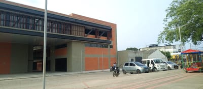 The image shows a street scene in the Perímetro Urbano Cúcuta, Colombia. The foreground is a paved plaza with red brick accents, leading to a street with parked cars and motorcycles. A building with a modern architectural style dominates the left side of the frame, featuring a combination of brick and concrete. Two people are riding a motorcycle, moving from left to right, close to a parked silver car. Further to the right, there is a white van, a white car, and a ball pit cart. The background shows buildings and a tree line under a lightly overcast sky.
