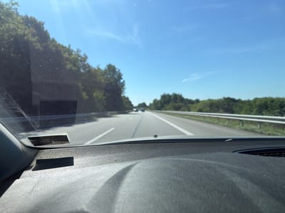 The image shows a view from inside a vehicle traveling on a highway. The perspective is from the driver's seat, looking forward through the windshield. The road ahead is a multi-lane highway with a white line marking the lanes. Trees and vegetation line the sides of the highway. The sky is clear and blue with a few wispy clouds. The sun is shining brightly, creating a glare on the windshield. The dashboard of the vehicle is visible in the foreground, with a dark, textured surface. There is a small sticker or label affixed to the windshield in the upper left corner. In the distance, other vehicles can be seen on the highway. The location is the city of Schönbek, Germany.