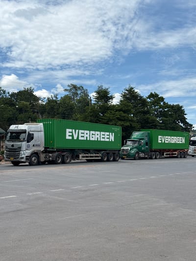 Two commercial trucks, each carrying a green shipping container labeled "EVERGREEN," are parked on a paved road. The truck on the left is silver and white, while the truck on the right is green. Trees and a blue sky with scattered clouds are visible in the background. The image appears to have been taken in Lao Bảo Commune, Vietnam, a town located near the border with Laos, known for its trade and transportation activities.
