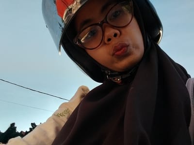 The media file is a close-up selfie of a young Indonesian woman wearing a black hijab and a dark brown hijab. She is also wearing a motorcycle helmet with a clear visor and red detailing. Her glasses have brown frames. She is looking towards the camera with her lips puckered. There is a faint embroidered green leaf pattern on her light-colored garment visible on the left side of the frame.

The scene appears to be outdoors, with a clear blue sky as the background, suggesting it is daytime. The presence of the motorcycle helmet and the woman's attire indicates she might be preparing for or has just finished a motorcycle ride. The photo was taken in Macege, Indonesia. The overall mood appears lighthearted and casual, given the selfie pose and facial expression. There is no visible text in the image.