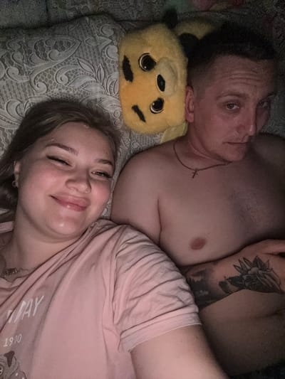 In an overhead shot, a young woman and a man are lying on a patterned bedspread. The woman is on the left, smiling and winking at the camera. She is wearing a pink t-shirt with text and a design of a dog on it, along with a necklace. The man is on the right, looking towards the camera with a serious expression. He is shirtless, wearing a necklace with a cross, and has tattoos on his arm. Between them is a yellow stuffed animal with large eyes and black markings. The lighting is dim, suggesting a nighttime setting. The location is identified as Sovkhoza "Razdol'e", Russia.
