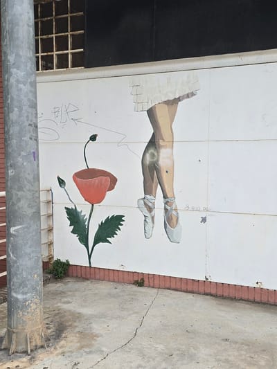 The image depicts a mural on a white wall in Cornellà de Llobregat, Spain. The mural features a drawing of a poppy flower with red petals and green leaves, as well as the lower legs and ballet shoes of a dancer. The dancer's legs are positioned crossed, and she is wearing white ballet slippers. There are faint graffiti markings and a signature that reads "S. RUIZ PALACIO ABDELL" on the wall. To the left of the mural is a weathered grey metal pole. In the background, above the mural, is a dark surface with a barred window. The ground in front of the wall is a concrete surface with some cracks and debris, and there are small green plants growing at the base of the wall.