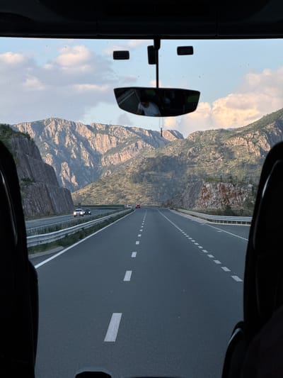 The scene shows a view from inside a vehicle, likely a car or bus, traveling on a multi-lane highway in Raći, Montenegro. A large, rocky mountain range rises to the left of the road. The highway has white lane markings and guardrails. In the distance, two vehicles are visible on the road. A rearview mirror is visible inside the vehicle, reflecting a person wearing a mask. The sky is partly cloudy. The image is taken from a low-angle perspective looking towards the horizon.