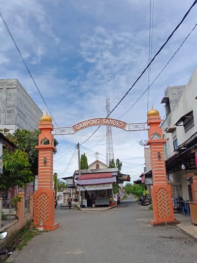 The image shows an archway with the text "GAMPONG SANGSO" in the city of Sangso, Indonesia. The archway is adorned with orange pillars topped with golden domes. To the left of the archway is a building with a concrete facade and some trees. To the right are buildings with shops and a telecommunications tower in the background. The road in front of the archway is paved and appears to be a street. There are a few people visible on the street, as well as some vehicles, including a motorcycle and a three-wheeled cart. The sky is cloudy and the overall scene suggests a typical day in a small Indonesian town.