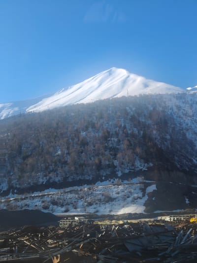 The image depicts a snow-covered mountain against a clear blue sky. The lower slopes are covered in trees, with patches of snow visible. A road or railway line cuts across the lower part of the mountain. In the foreground, there is a debris field, including metal objects and remnants of buildings. A building with a partially standing frame is also visible within the debris. The location is Almasiani, Georgia.