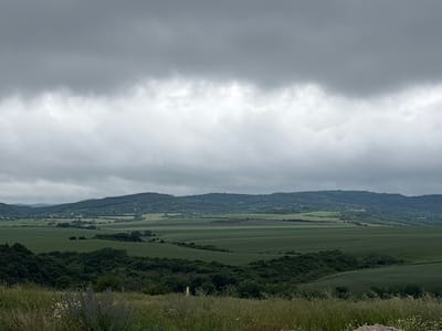 This image depicts a panoramic view of rolling hills and agricultural fields under a heavily overcast sky. The foreground features tall grasses and wildflowers, while the midground showcases lush green fields with some patches of darker vegetation. In the distance, a series of tree-covered hills forms a mountain range. A small village or cluster of buildings can be seen nestled in a valley between the fields and the distant hills. The scene is characteristic of the rural landscape surrounding Slivnitsa, Bulgaria, which is known for its agricultural plains and hilly terrain. The overall atmosphere is subdued due to the cloudy weather.