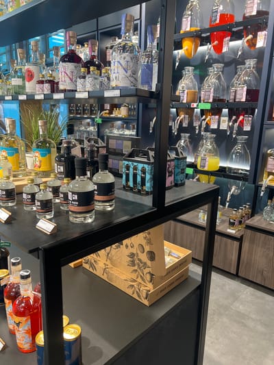 This is a vertical, eye-level shot of a retail display inside a store, likely a liquor store or a specialty shop. The display features a large selection of bottles, primarily gin, arranged on shelves. The bottles vary in size, color, and design, with some containing clear liquids and others colored spirits. Many bottles have distinct labels with text, including brand names like "SMASH GIN," "BOTANICAL," and "BANCAL GIN." Prices are visible on some labels, such as "25.95€" and "15,95€."
In the background, glass dispensers with various colored liquids are visible, as well as shelves stocked with more bottles. Some boxed items, possibly gift sets or packaging for the spirits, are also present. The overall aesthetic is modern and clean, with dark shelving and display cases. The lighting suggests an indoor environment, likely during daytime hours, given the brightness of the store. No people are visible in the image. The context suggests a commercial setting in Aachen, Germany, where these spirits are being offered for sale. The prominent display of gin suggests a focus on this particular type of alcoholic beverage.