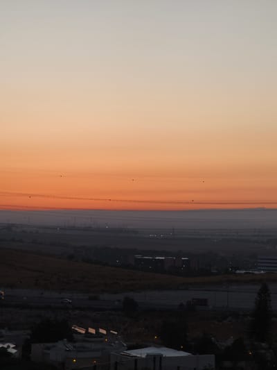 This image depicts a wide, horizontal landscape scene at dusk, featuring a gradient sky transitioning from light grey at the top to a vibrant orange near the horizon. A series of power lines crisscross the sky, with small, dark circular insulators visible on them. Below the sky, a hazy band of lighter grey stretches across the midground, obscuring the distant landscape. In the foreground, the scene transitions to a darker, silhouetted environment. On the left, a hilly, dry, golden-brown landscape is visible, leading to a residential area with low-rise buildings. Several of these buildings have solar panels installed on their roofs, reflecting the dim light. A white SUV is driving on a road in the mid-left of the frame, and a red truck is visible further to the right on a road in the darker foreground. The overall impression is a panoramic view of the outskirts of Be'er-Sheva, Israel, as daylight fades.
