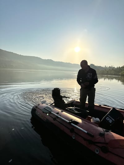 A man stands in an inflatable boat on a calm body of water at sunrise. The sun is low in the sky, casting a warm glow and creating lens flare. The man is wearing a dark hoodie with text and khaki pants. He appears to be looking down at something in the boat, which contains a Mercury outboard motor with "50" prominently displayed. There are also oars inside the boat. In the background, misty hills rise from the water, covered in trees. The overall scene suggests a peaceful start to the day, possibly for fishing or some other water activity. The image has been rotated 90 degrees clockwise.