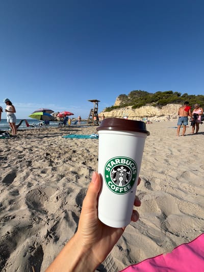 A first-person perspective shot shows a hand holding a white Starbucks coffee cup with its logo visible, set against a sandy beach with the ocean and a rocky, tree-covered hillside in the background. The sky is clear blue. In the mid-ground, several beach umbrellas, chairs, and people are scattered on the sand. A lifeguard tower is visible on the right side of the frame. The overall scene depicts a sunny day at the beach in Urbanització Cales I Talaies, Spain.