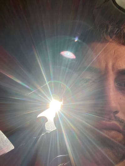 The image is a close-up, overexposed photograph of a man's face, taken from a low angle. The sun is directly in front of the camera, creating a strong lens flare that obscures much of the scene. The man appears to be Caucasian, with short, dark hair and stubble on his face. He is looking towards the camera, and his right eye is visible. The background is dark, with some indistinct shapes that may be part of a car interior. The image does not contain any information about the city of Rogosh, Bulgaria.