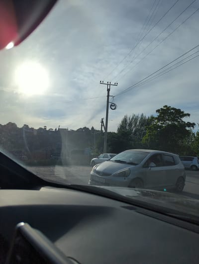 The image shows a view from inside a car on a road lined with power lines and utility poles. The sun is low in the sky. Adjacent to the road is vegetation. In the foreground is the interior of the car. Parked cars are visible next to the subject car. The location is believed to be Beş-Küŋgöy, Kyrgyzstan, inferred from the license plates of the cars. A hillside with buildings is also visible further in the background.