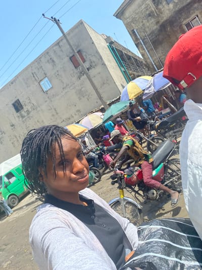 The image captures a vibrant and bustling street scene, likely a market or commercial area, under bright daylight. In the immediate foreground, a person with dark braided hair and dark skin is taking a selfie, looking directly at the camera with a neutral expression. They are wearing a light-colored, possibly white or grey, long-sleeved top over a dark vest. Partially visible above them is another individual wearing a red baseball cap.
The background is a dynamic tableau of daily life. Numerous motorcycles, suggesting a reliance on "okada" (motorcycle taxi) transport, are scattered throughout the scene, some parked and others with people interacting with them. Several market stalls are identifiable by colorful umbrellas—one yellow and blue, another light blue—providing shade for vendors or customers. People are seen milling about, with one individual prominently wearing a yellow, green, and black patterned shirt near a motorcycle. The ground appears uneven, a mix of dirt and possibly some pavement, with patches that might be muddy or dusty.
Multi-story concrete buildings, showing signs of wear and age with visible windows and utility fixtures, line the street under a clear, bright blue sky. Utility poles with wires are also visible, running across the upper part of the image. A green vehicle, possibly a minivan or small bus, is partially visible in the lower right background. The strong sunlight casts clear shadows, indicating it is daytime. No discernible text is clearly visible on any signs or objects. The overall scene suggests a busy urban environment in Satellite Town, Nigeria.