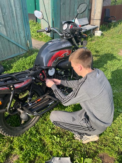 In Nizhnii Bugrysh, Russia, a young male, dressed in a grey Adidas tracksuit and athletic shoes, kneels on a grassy area in front of a motorcycle. He is actively working on the motorcycle's electrical components, his hands engaged with wiring. A toolkit containing an orange-handled screwdriver and a wrench is placed on the motorcycle's rear rack. The background features a weathered green wooden fence and a glimpse of a rural setting with overgrown grass, a bench, a chair, and a white bucket. The scene is illuminated by bright sunlight, casting distinct shadows.