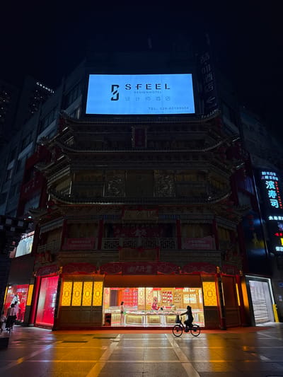 The image is an eye-level, nighttime exterior shot of a street in the Jinjiang District of Chengdu, China. Prominent is a multi-tiered, ornate building in a traditional Chinese architectural style, illuminated with golden lights. Adjacent to it is a large, vertical sign displaying "SFEEL DESIGNHOTEL" with its logo, and a phone number: TEL: 028-80199904. A smaller sign above directs to "喆・啡酒店" (Zhe. Fei Hotel) "turn left 20 meters".
To the right, the lower levels of the building feature brightly lit storefronts, likely jewelry shops. A person on a bicycle is silhouetted in one entrance, and a few pedestrians are walking on the sidewalk in front. Other signs are visible, including one for "HAMA-SUSHI" (濱寿司), with others for “阳光百货” (Sunshine Department Store) and “成都急店” (Chengdu Fast Shop). A taller, modern building is partially visible on the left of the frame.
