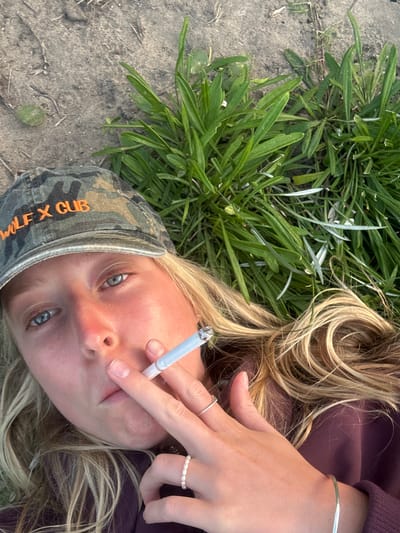 A young woman is lying on the ground, possibly in a park in Wollongong City Council, Australia. She is blonde and wearing a camouflage cap with "WILD X CUB" written on it in orange. She is holding a lit cigarette to her lips. She has a silver bracelet on her right wrist and rings on her right hand. She is wearing a dark-colored top. She is surrounded by grass and dirt. The image is taken from an overhead perspective.
