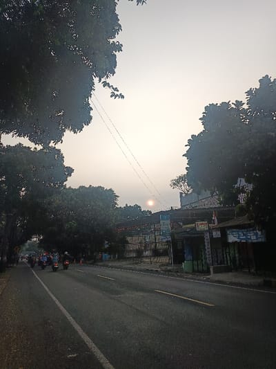 An image captured in Muka, Indonesia, depicts a roadside scene under a hazy sky with the sun appearing as a distinct orange-yellow disc in the upper mid-right. The perspective is from the roadside, looking along a paved asphalt road. The road features a solid white line on its left edge and a dashed yellow line separating lanes. On the left side of the road, a column of at least five motorcycles, each operated by a helmeted rider, travels away from the viewer. Dense green tree foliage lines the left side of the road, creating a partial canopy, and also frames the upper right portion of the sky. On the right side of the road, a series of low-rise commercial structures are visible. These buildings display multiple signs, including a blue sign with "ATM" and a logo, and another sign indicating "Canon PHOTO COPY." An Indonesian flag is mounted on one of the structures. Multiple thin utility lines traverse the upper portion of the frame. The scene is illuminated by the low-angle sun, suggesting either early morning or late afternoon.