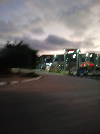 A hazy, vertically-oriented image depicts a cityscape beside a grey surface. Buildings are visible with illuminated signs and windows. In the foreground, blurred, cloud-like formations obscure some details. Some greenery is also present. The overall scene suggests an urban environment at night, possibly with a weather phenomenon occurring.
